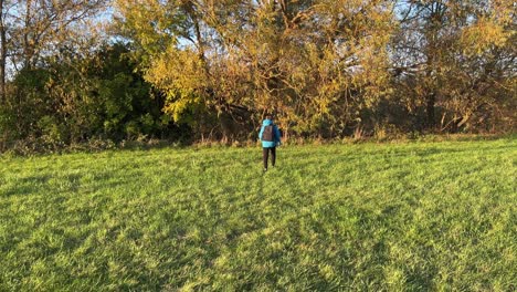 Young-boy-walking-away-with-rucksack-towards-trees-in-a-field