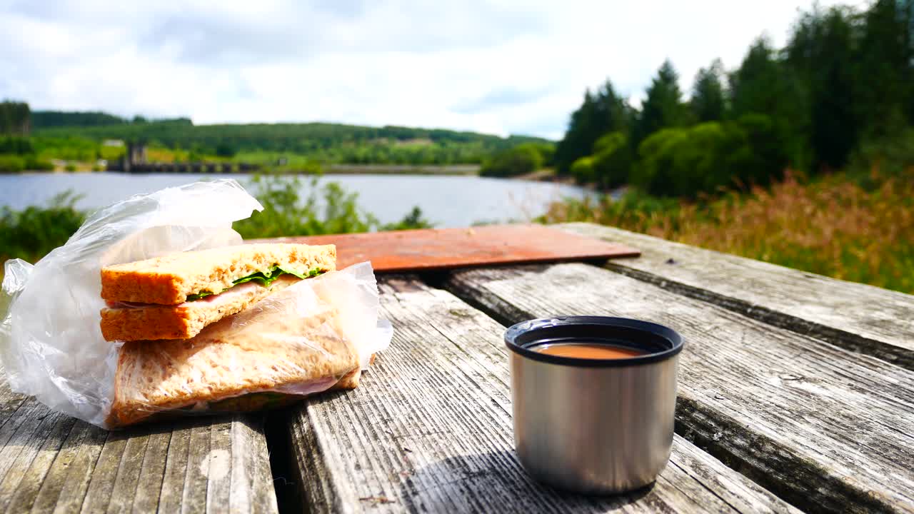 relajarse en una mesa de picnic de madera con un matraz de té y un sándwich de ensalada de jamón con vistas al soleado paisaje del lago azul del bosque