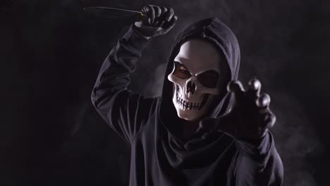 close up of scary man in the hooded sweatshirt wearing halloween mask holding a knife in the threatening gesture, a symbol of murder and crime, on the black background with smoke