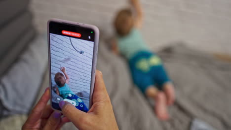 mommy is taking video of her son on the smartphone. little baby boy crawling by the bed exploring the white brick wall. blurred backdrop.