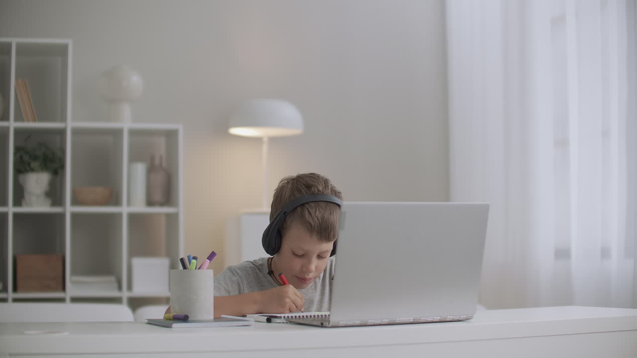 un niño está viendo videos educativos para niños en una computadora portátil y escuchando con auriculares dibujando en papel