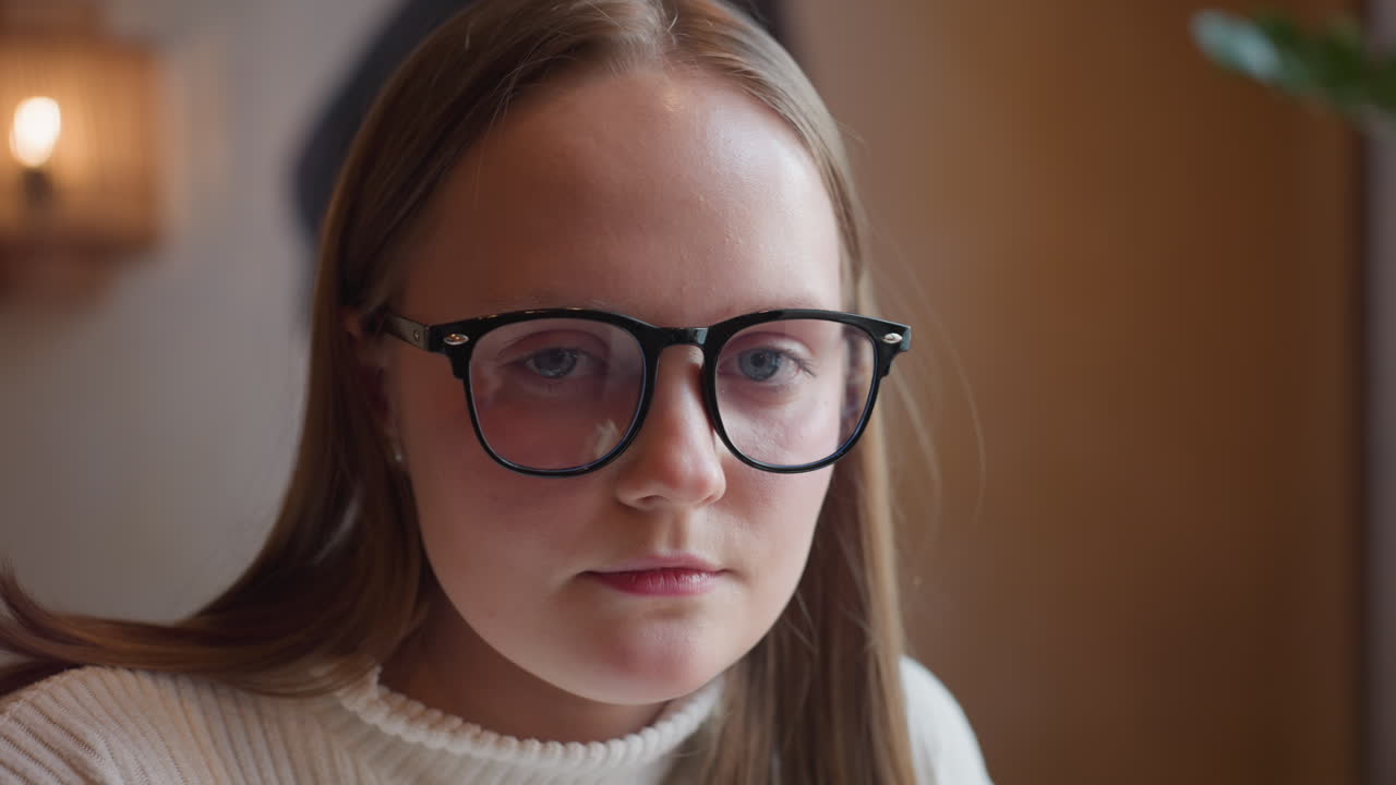 young girl in glasses wearing white ribbed sweater gazes seriously at camera while seated indoors, soft wall lighting and neutral background