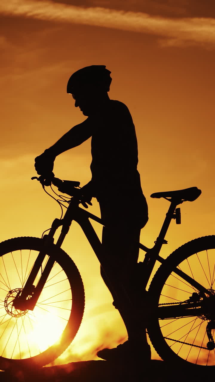 Silhouette of a biker with his bike walks outdoors at sunset. Healthy cyclist walking in the evening and a man's silhouette going by against the beautiful summer sunset. Vertical video
