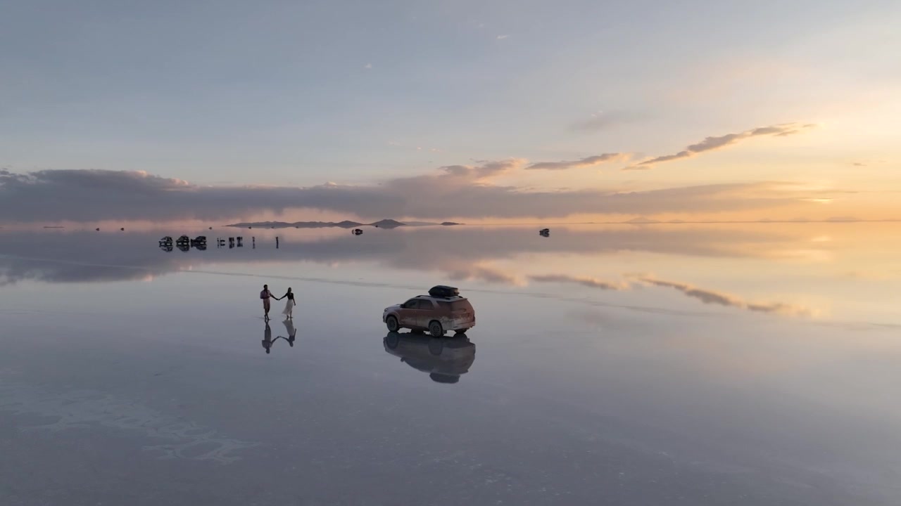 Salar de Uyuni ao pôr do sol com uma pessoa caminhando perto de um veículo estacionado, criando um reflexo espelhado na superfície da água, Bolívia em vista aérea
