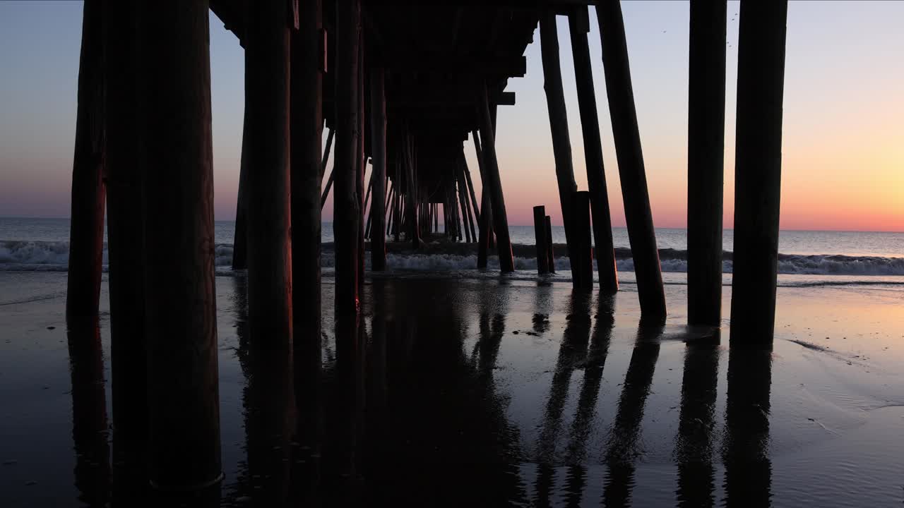 Slow motion sunrise view of waves rolling in and crashing against the wooden supports of a fishing pier on a beach