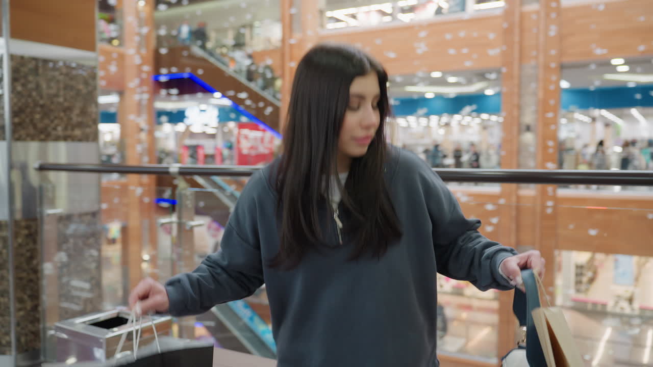 Young woman in loose sweatshirt walks inside modern shopping mall after completing shopping, looking around while waiting for friend, surrounded by glass railing and colorful background