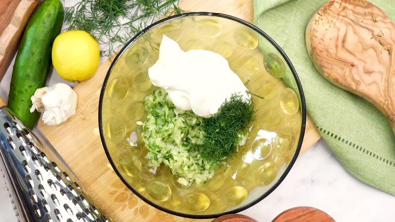 Making Tzatziki Greek or Mediterranean sauce or dip for a party. Dip or sauce in a green bowl on a wooden cutting board with dill, green napkin and a grater. Adding kosher salt into the Tzatziki sauce