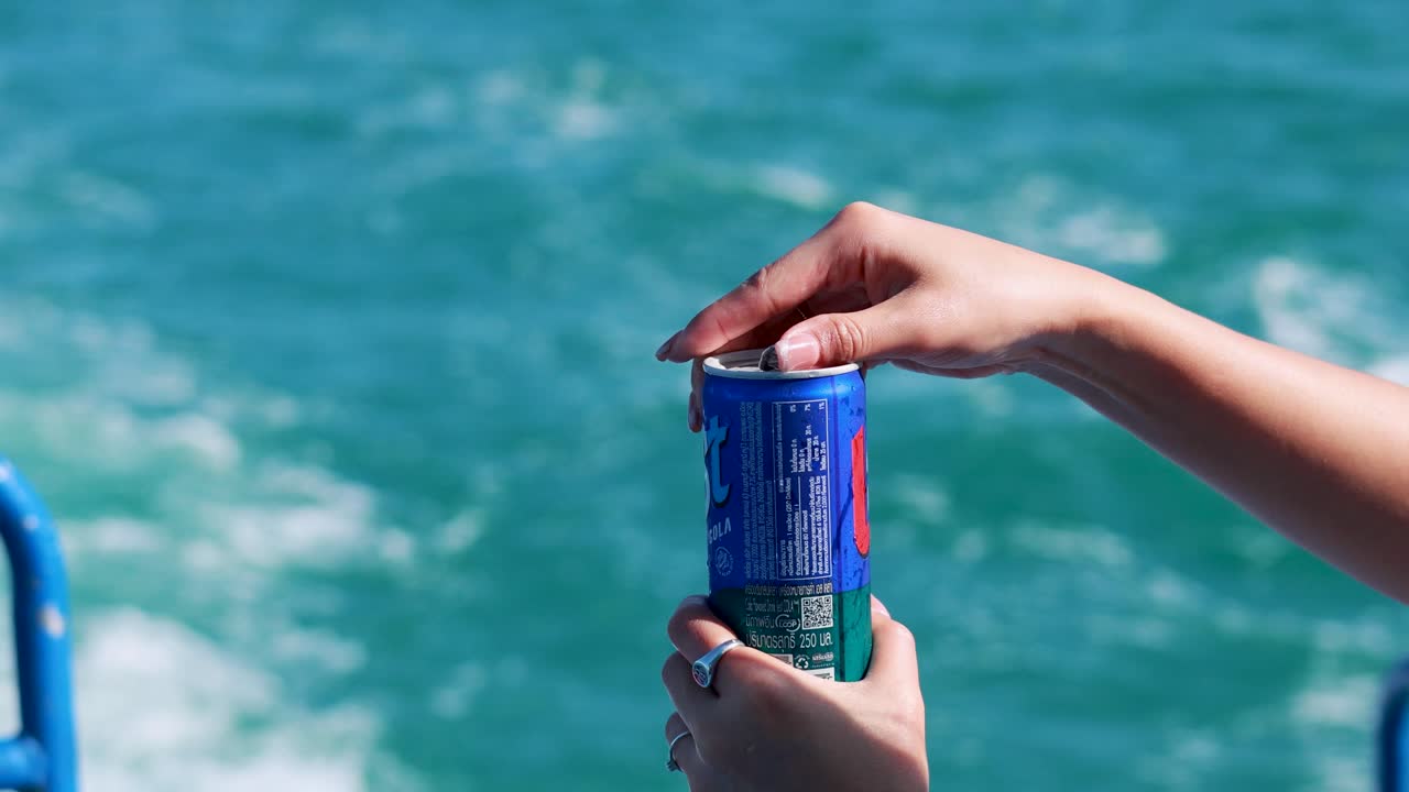 A person opens a soda can on a boat with vibrant ocean waves in the background under bright sunlight