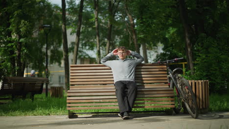 boy wearing glasses, gray top, and black trousers sits on wooden bench in outdoor park, stretching and adjusting posture while resting, parked bicycle beside him, surrounded by green trees