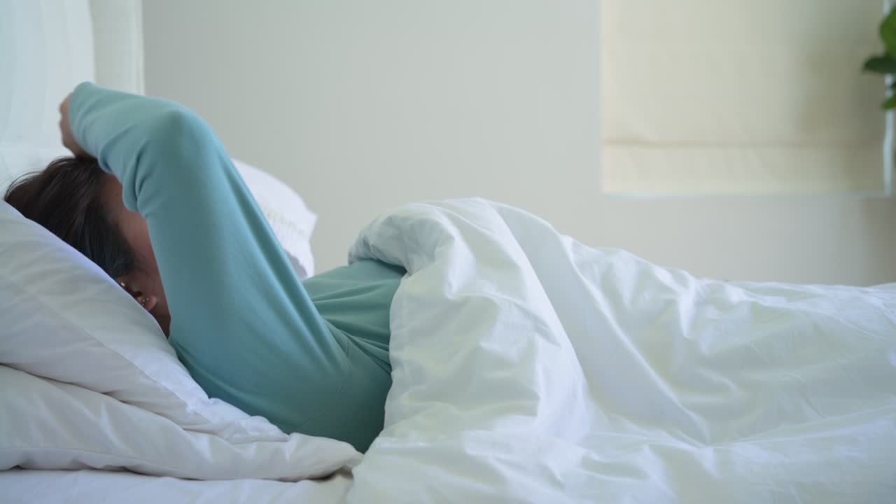 Donna che si sveglia in una camera da letto luminosa, si stira, poi tira via il piumone dal letto per alzarsi