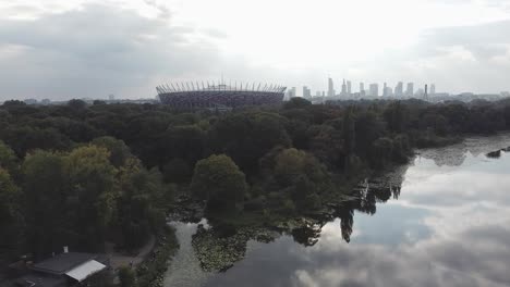 Toma-Aérea-De-Drones-Del-Estadio-De-Varsovia,-Con-Efecto-De-Paralaje-Con-El-Horizonte-De-La-Ciudad,-El-Lago-Y-El-Bosque-En-El-Fondo