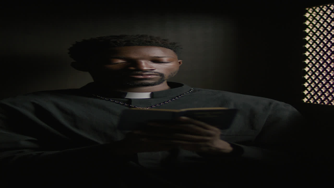 African American Priest Holding Bible and Reading Scripture in Confessional