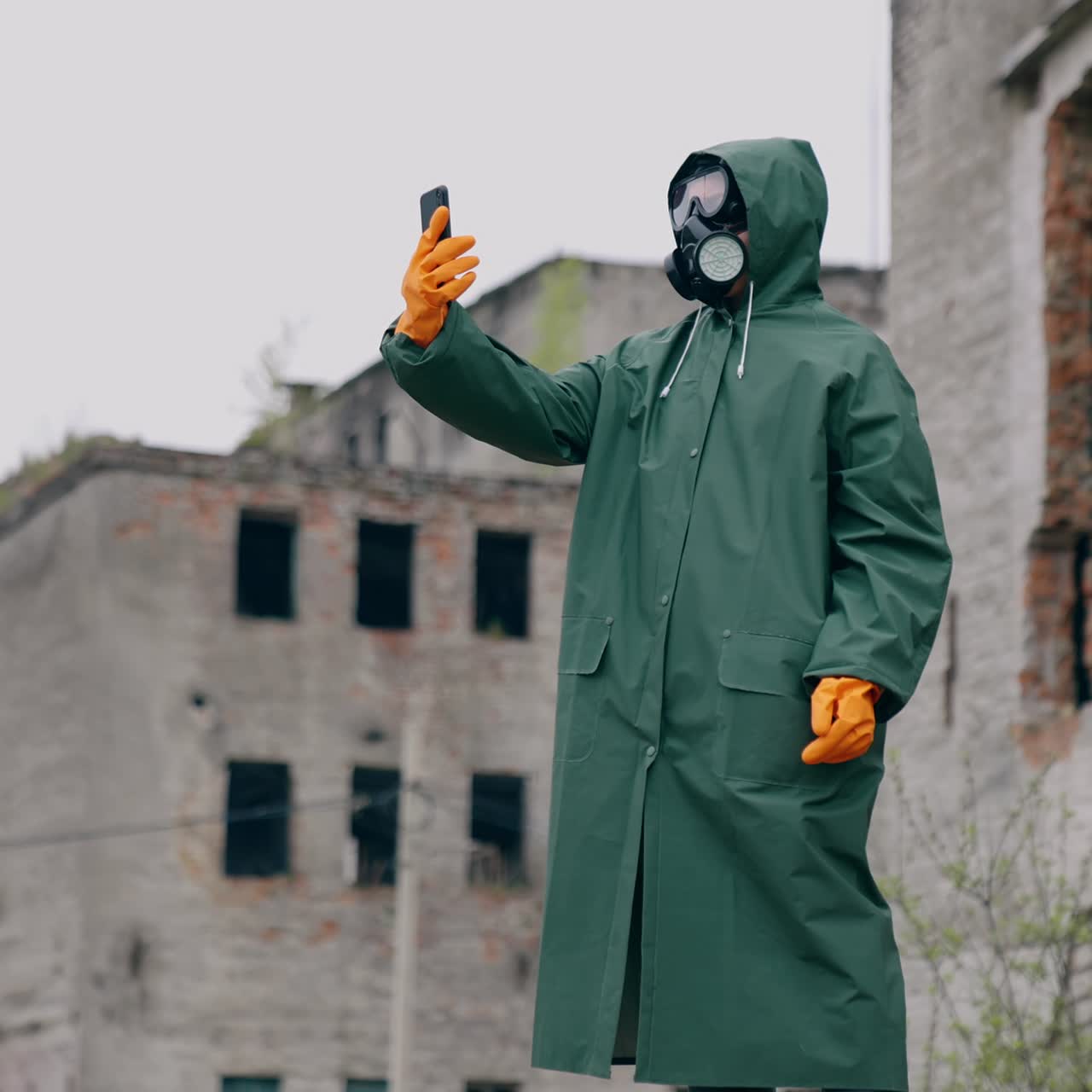 Human in a protective suit and gas mask with a phone. Unrecognizable person in safety uniform, gloves and respirator doing selfie in the destroyed place during radiation
