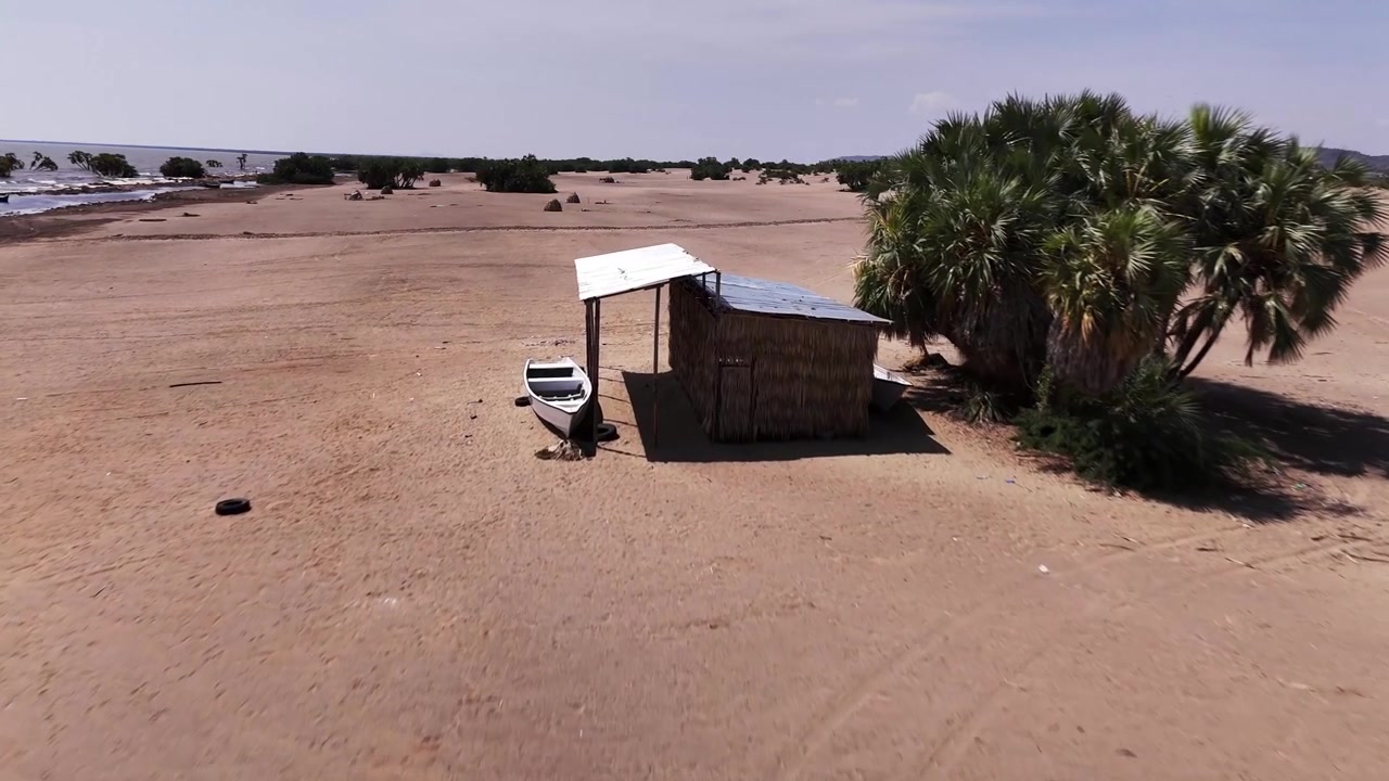 Aerial view of a lakeside shelter and small boat on the sandy shore of Lake Turkana, Kenya, surrounded by doum palms and arid terrain.