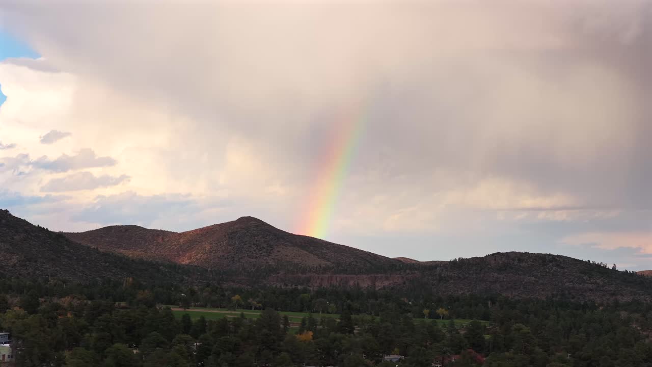 Aerial slide revealing a soft rainbow forming over a mountain ridge under stormy skies. Scenic, atmospheric, and ideal for nature, travel, or wellness visuals
