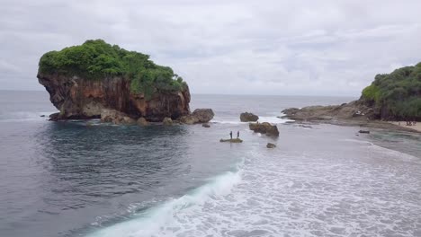 aerial view of two people standing on rock in bay of jungwok beach in indonesia during cloudy day - giant boulder with growing trees in background