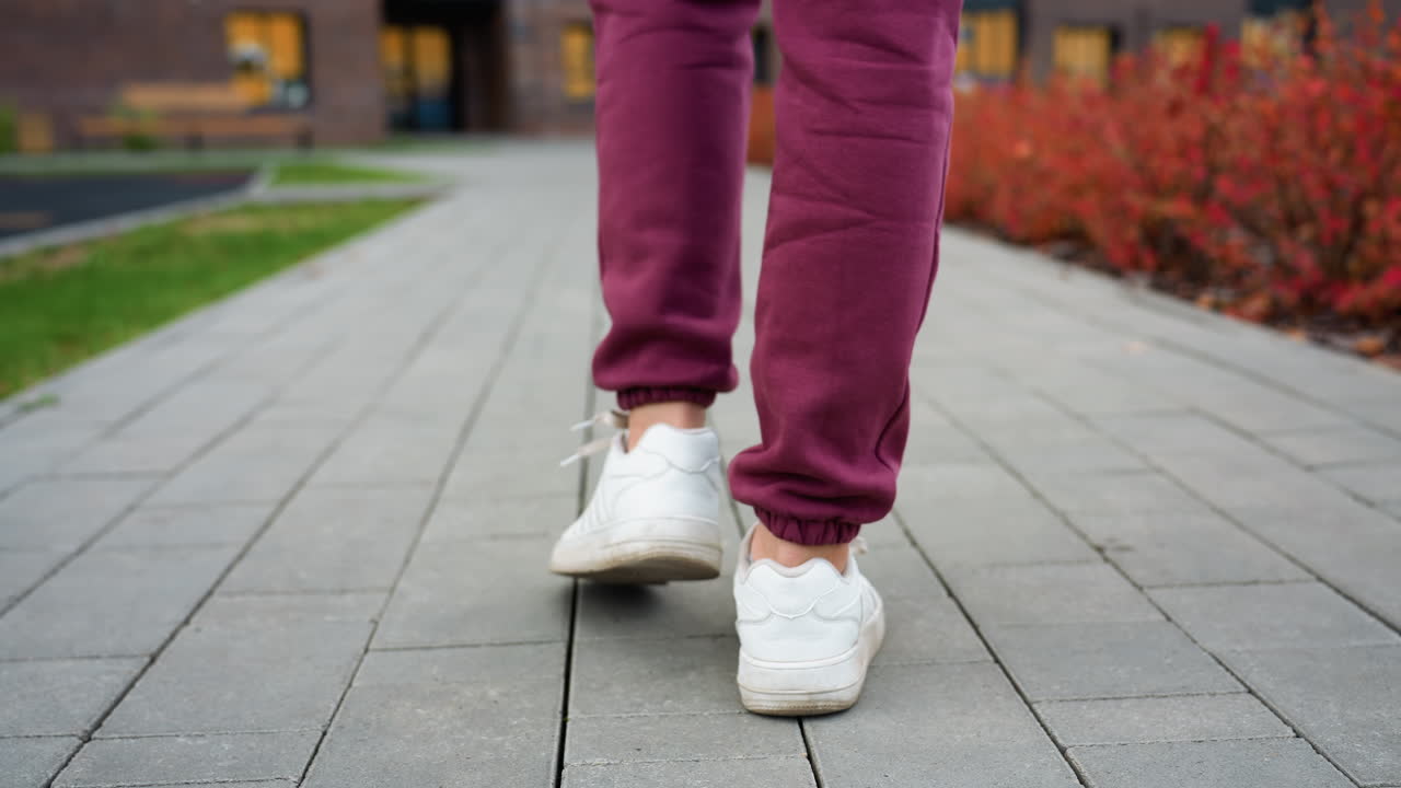 Leg view of gym enthusiast wearing white sneakers stepping on grey tiled pavement alongside red spring shrubs in urban outdoor fitness area showcasing stride motion in morning light