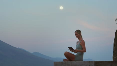 woman sitting in easy pose looking at full moon and writing mental note in journal