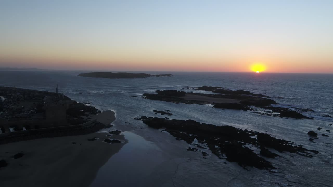Droneoptagelse i fugleperspektiv, der bevæger sig baglæns (trækker tilbage) for at afsløre den robuste, stenede kystlinje og havets bølger ved solnedgang. Kameraet trækker sig væk fra solen og udvider udsigten over de dramatiske klipper