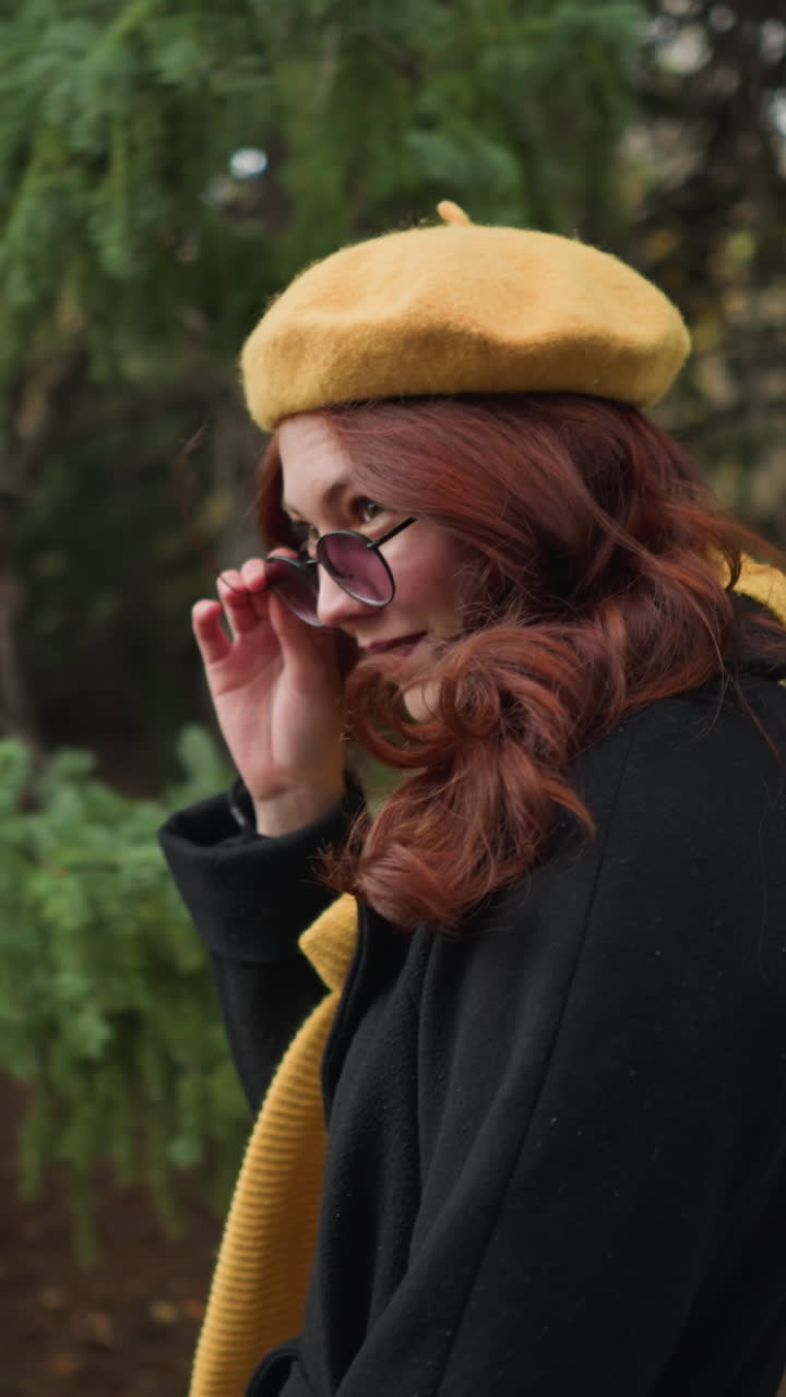 Donna universitaria con lunghi capelli rossi ondulati, che indossa un cappotto nero e un basco giallo con sciarpa abbinata, si sistema gli occhiali da sole mentre guarda in alto, camminando attraverso un parco verde con un colorato fogliame autunnale.