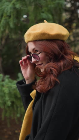 dama de la universidad con largo cabello rojo ondulado, con un abrigo negro y boina amarilla con pañuelo a juego, ajusta sus gafas de sol mientras mira hacia arriba, caminando a través de un parque verde con colorido follaje de otoño