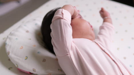 acostada en un colchón y su cabeza en una almohada, una bebé recién nacida está estirando sus brazos y piernas mientras se acaricia la cara