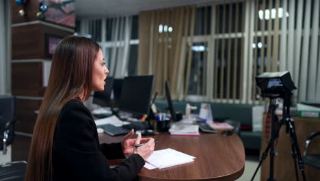 brunette long-haired woman sits at desk talking. side view. shooting a video for blog in the office.