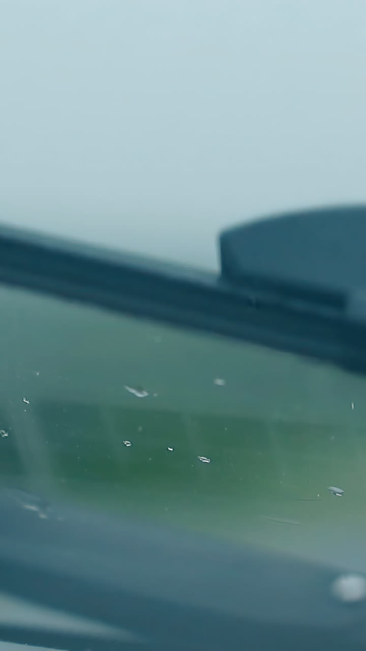 close view from car cabin black metal wiper works under light rain against green blurry silhouette