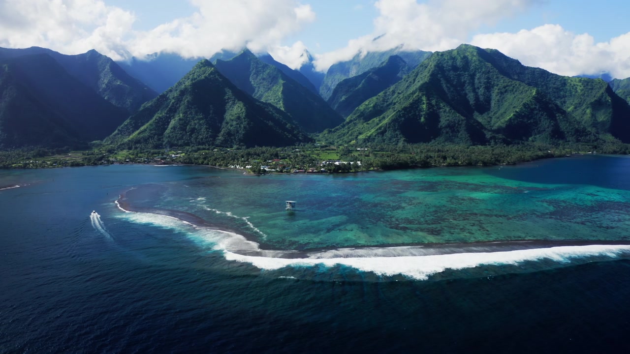 Dronen ilmakuva Teahupoon kylästä, Tahitilta, aalto murtuu avomerellä koralliriutalla sademetsävuorten noustessa talojen takana.
