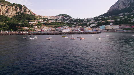 An-extreme-wide-drone-shot-of-boats-in-the-sea-around-the-tourist-port-of-Marina-Grande-on-the-island-of-Capri-in-Campania,-Italy,-with-a-lush-island-backdrop,-rocky-cliffs-and-pastel-buildings