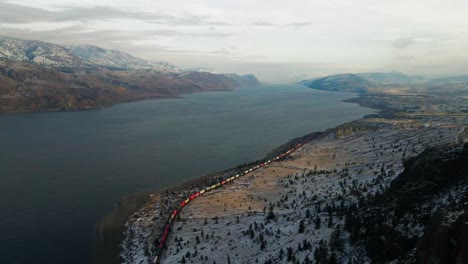 on a sunny winter's day, a colorful train drives along the shores of kamloops lake's hilly desert landscape, partially covered with snow