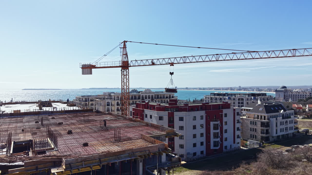 Lavori di costruzione in un cantiere vicino all'acqua durante una giornata di sole, con una gru che solleva materiali sopra le strutture vicine