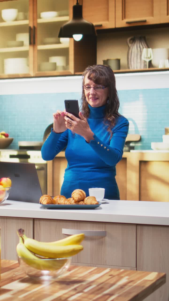 Vertical Video Elderly woman scrolling on smartphone apps by the counter in kitchen