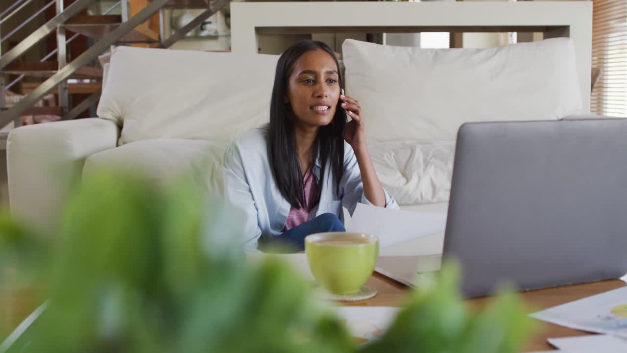 Mixed race woman using laptop talking on phone drinking coffee working from home