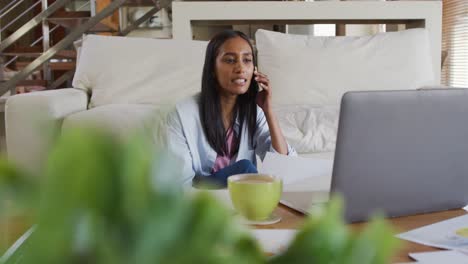 mixed race woman using laptop talking on phone drinking coffee working from home