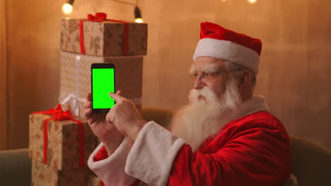 Santa-Claus-is-sitting-on-the-sofa-in-the-background-of-a-Christmas-tree-and-garlands-holding-a-mobile-phone-with-a-green-screen-pointing-at-it-with-his-finger.-Santa-holds-a-phone-with-a-chromakey-on-the-screen