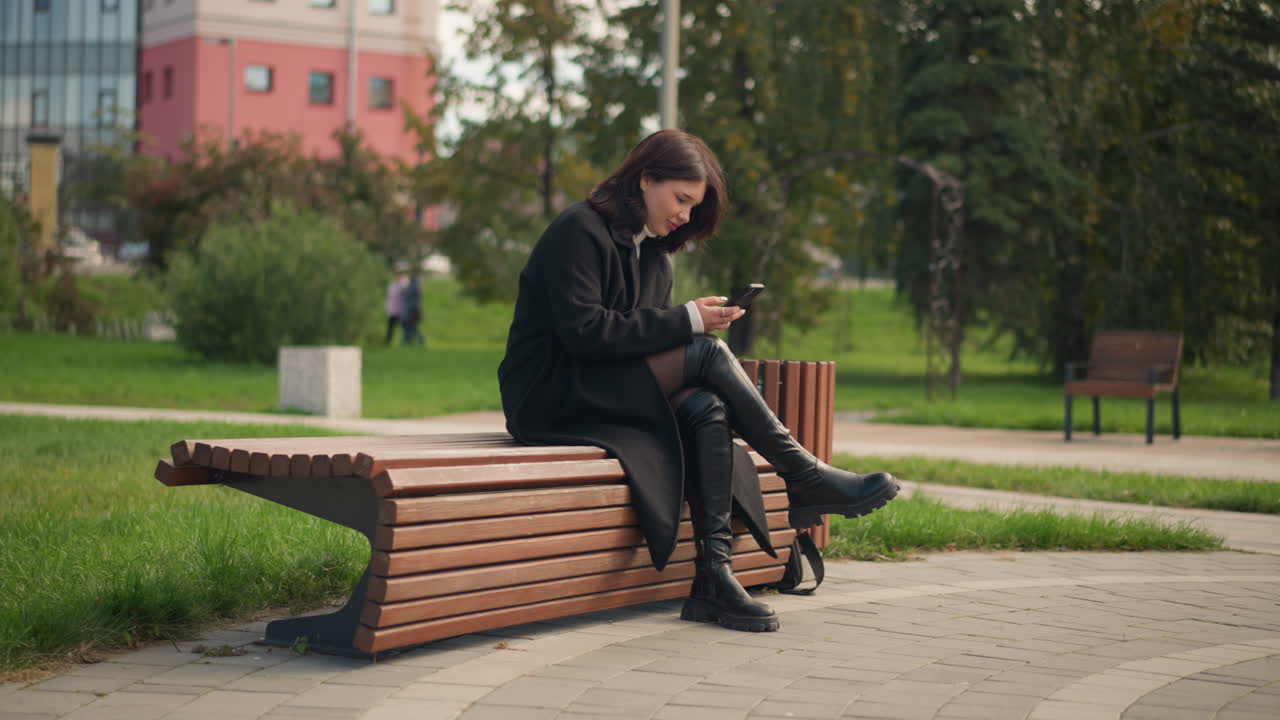 mujer sentada en un banco del parque usando un teléfono inteligente, vestida con abrigo y botas negras, rodeada de árboles verdes exuberantes, en un parque urbano con una vista borrosa de personas caminando en el fondo