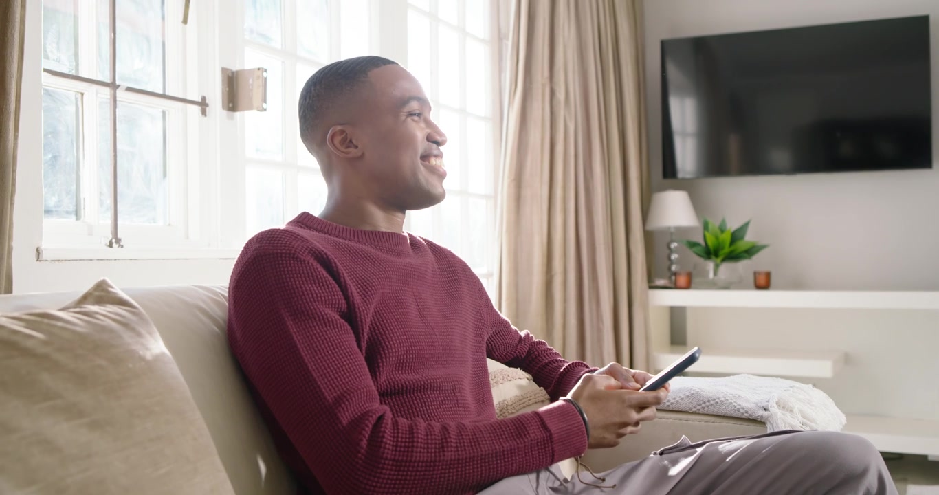 African American man sitting on sofa holding phone, tapping and reading as sunlight streaming