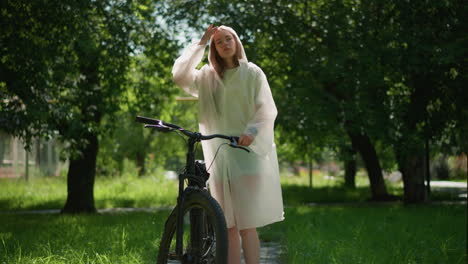 young lady in translucent raincoat pauses along sunny path with bicycle, placing stand near pole and removing hood to lean thoughtfully on handlebars, hand on chin, with greenery background