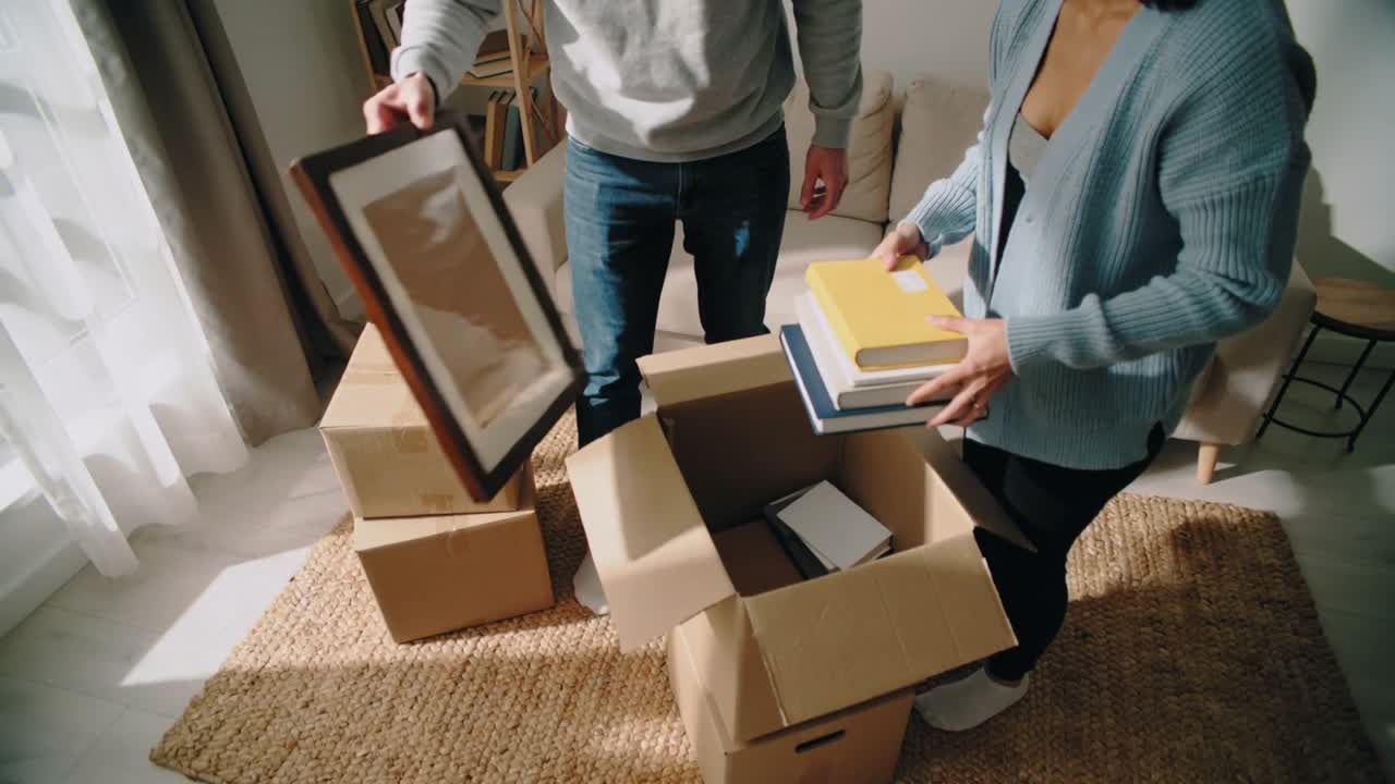 Couple reaching into open box, unpacking framed picture and yellow book, arranging for new home