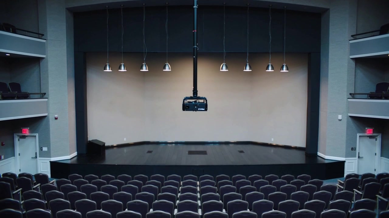 Luci sceniche a sospensione che si accendono, l'area del palco illumina lentamente una parete beige dal centro posteriore.