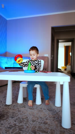 happy toddler sitting at desk plays with toy pyramid. smiling baby playing peacefully at home. vertical video.