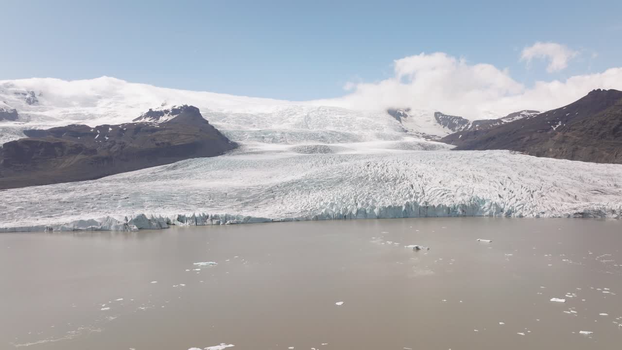 Wide view of Fjallsjökull Glacier descending into Fjallsarlon Lagoon, Iceland, with snow-covered peaks and tranquil waters under a bright blue sky.