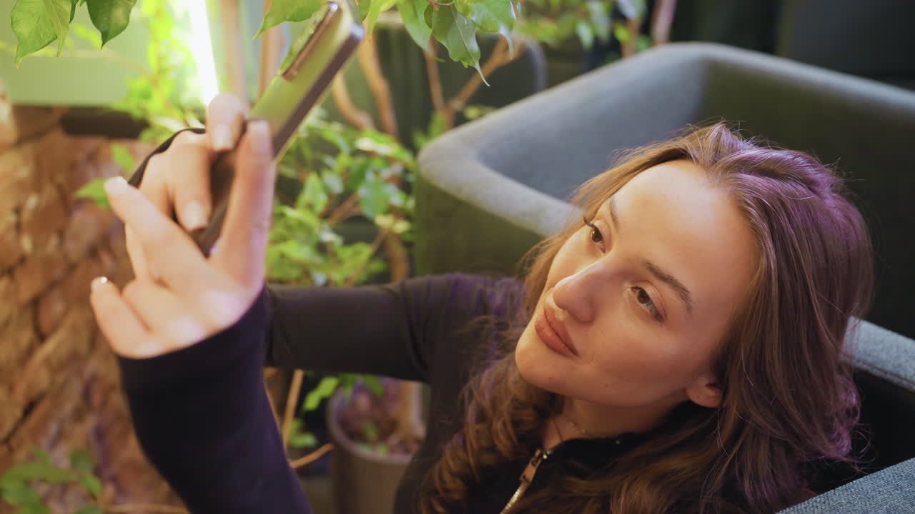 Lady with brown hair lifts phone while sitting beside green plants and soft chair, surrounded by warm natural light and cozy brick wall backdrop, immersed in relaxed moment with closed eyes