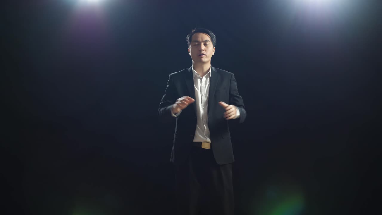 비즈니스 슈트를 입은 아시아인 스피커가 검은 스크린 스튜디오에서 말하는 동안 손을 함께 잡고 지표손가락을 나타니다.