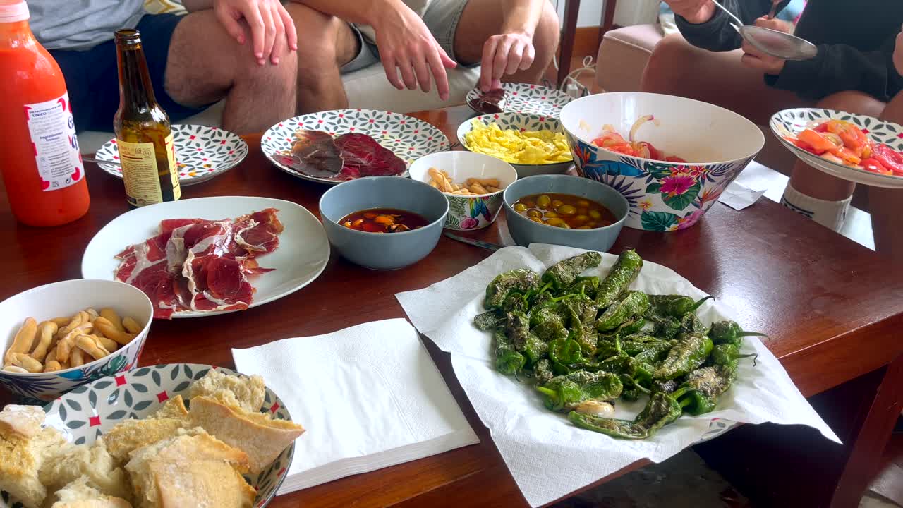 Group of friends enjoying tapas including ham, padrón peppers, and tomato salad