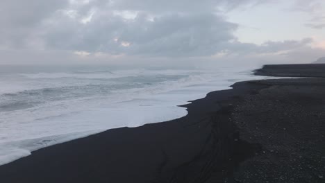 Aerial-landscape-view-of-ocean-waves-crashing-on-Iceland-Sólheimasandur-black-sand-beach,-on-a-cloudy-evening