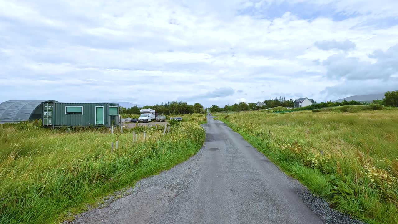 A vehicle travels along a narrow country road bordered by grassy fields and farm buildings under overcast daylight, with steady forward camera movement