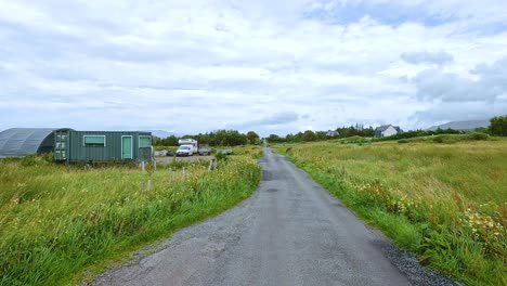 a vehicle travels along a narrow country road bordered by grassy fields and farm buildings under overcast daylight, with steady forward camera movement