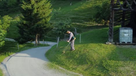 man in working outfit, mowing lawn with hand held nylon rotary petrol mower around his barn